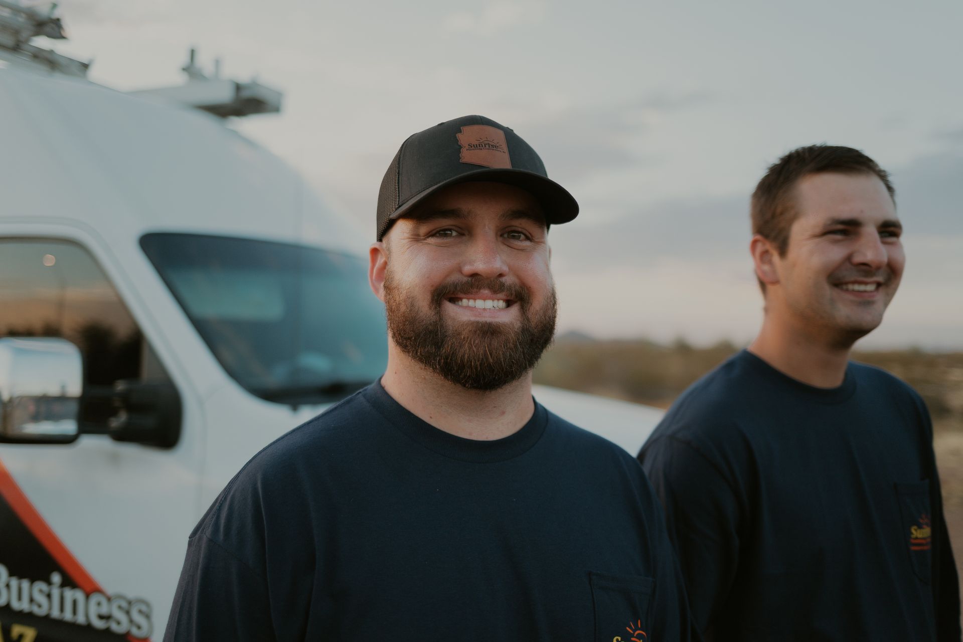 Two men smile in front of a white work van. One wears a hat, both have dark blue shirts.