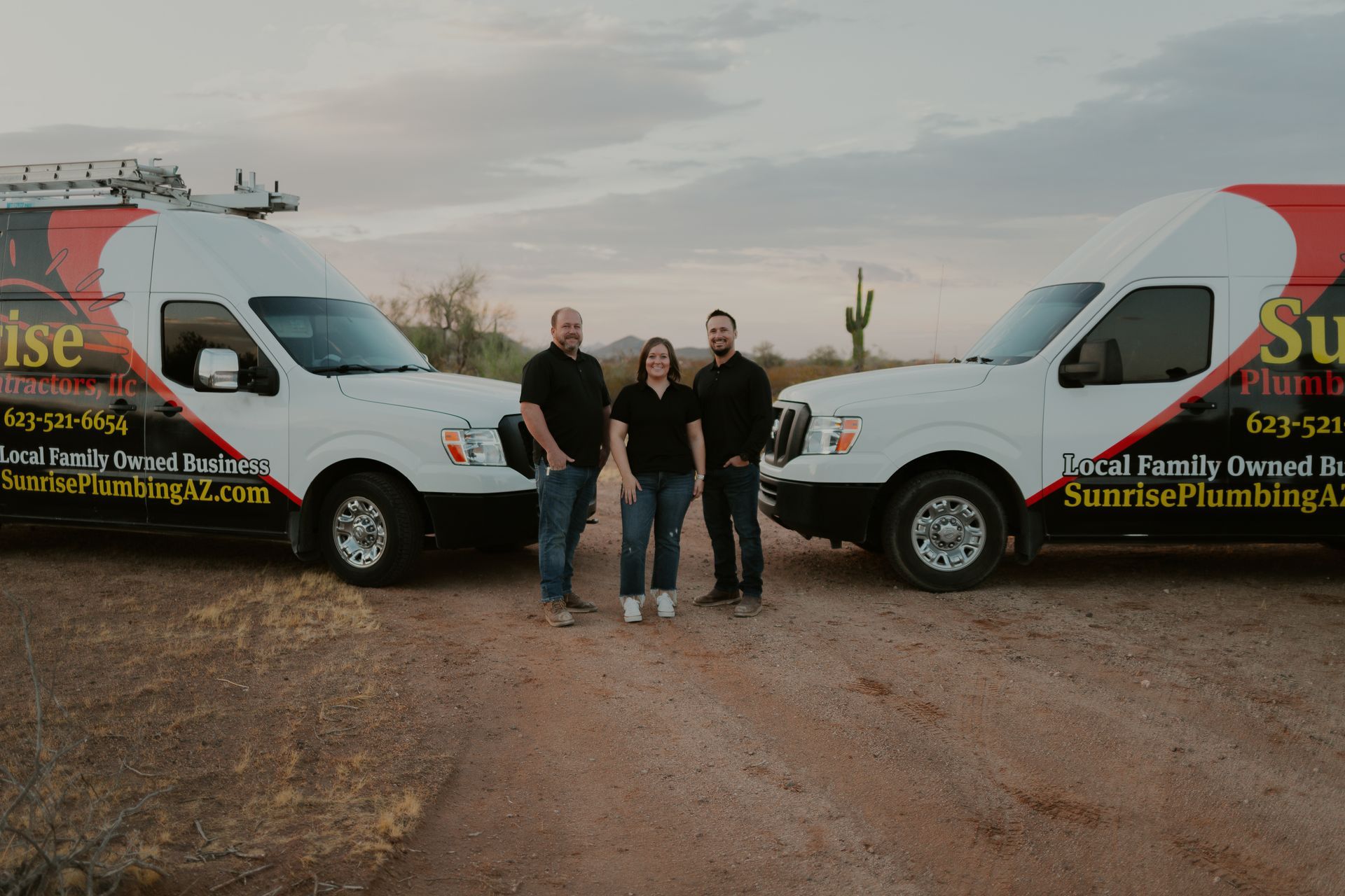 Three people stand between two Sunrise Plumbing vans in a desert landscape.