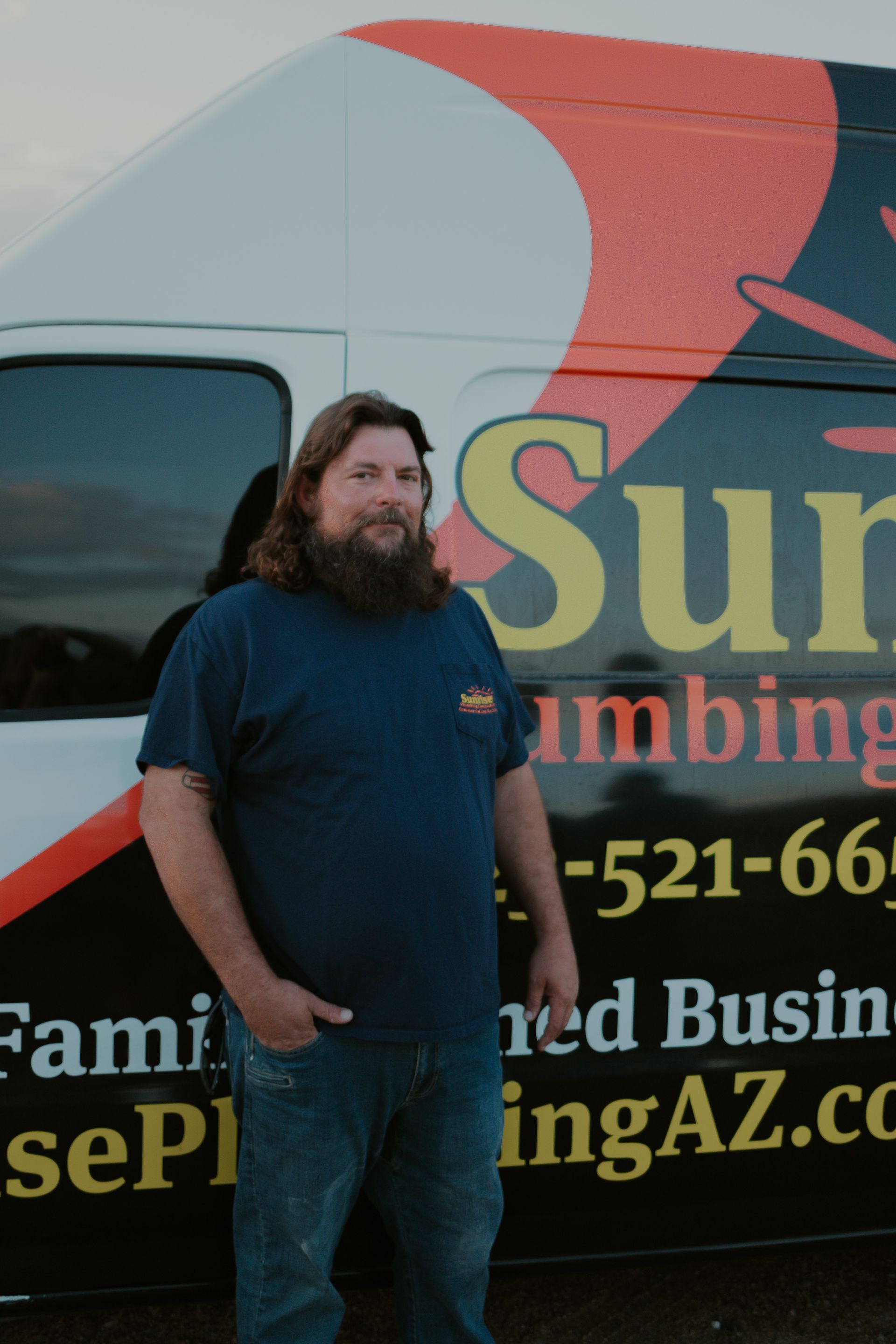 Man with beard stands beside a plumbing van with business logo.