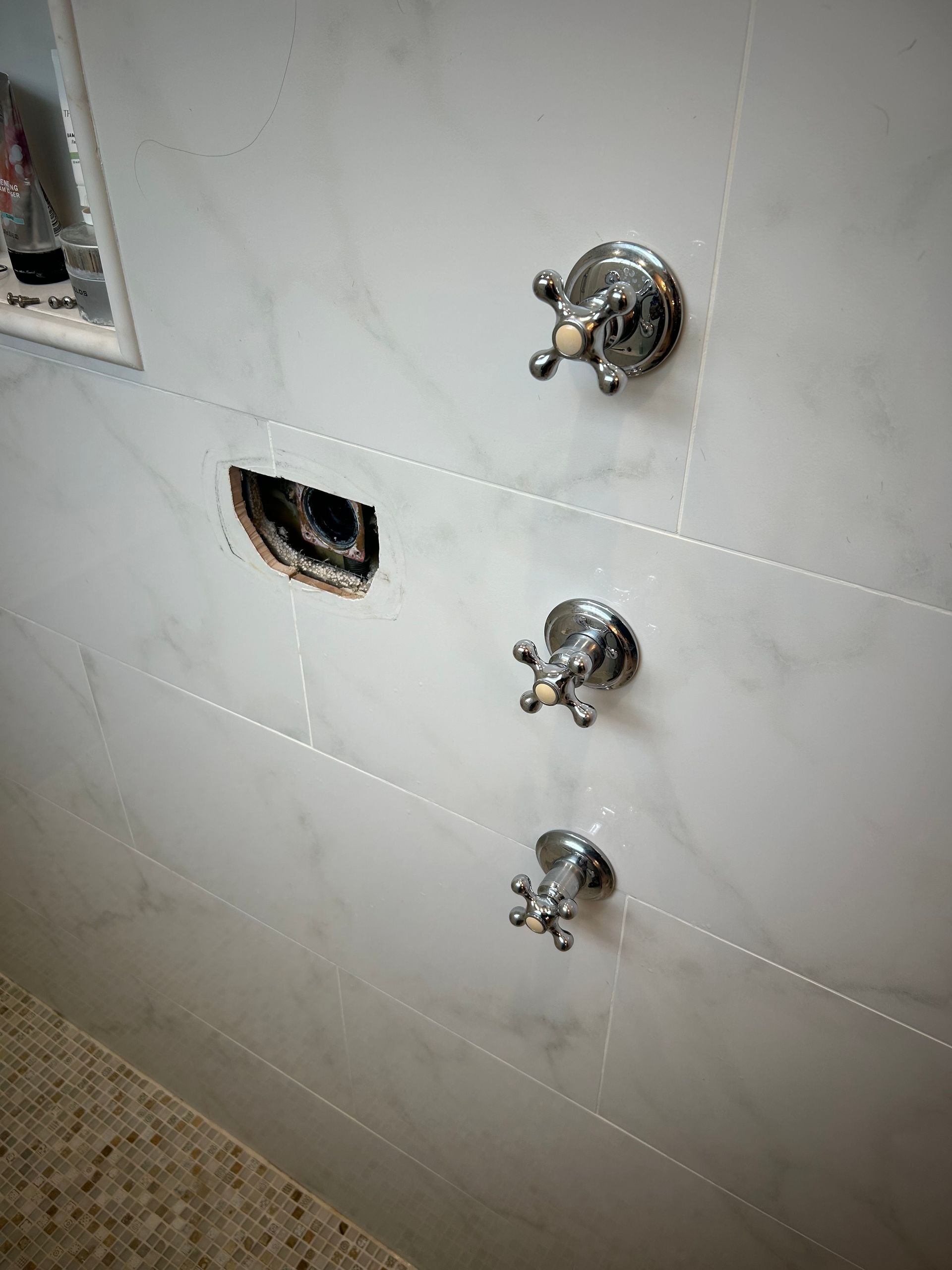 White tiled shower wall with three chrome faucet controls and an opening in the tiles.