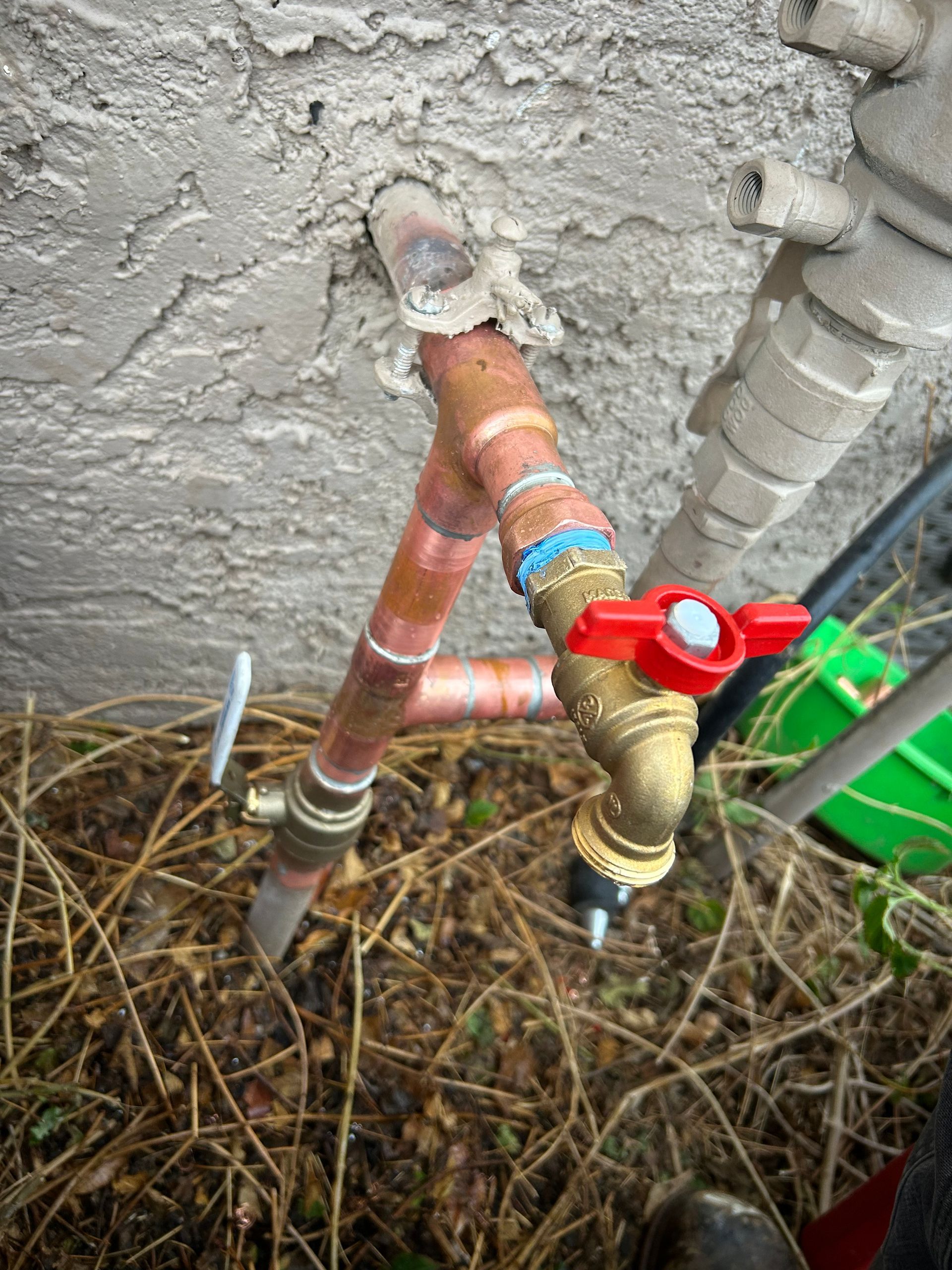Copper pipe water spigot, installed on a stucco wall. Red valve handle is open. Brown grass in foreground.