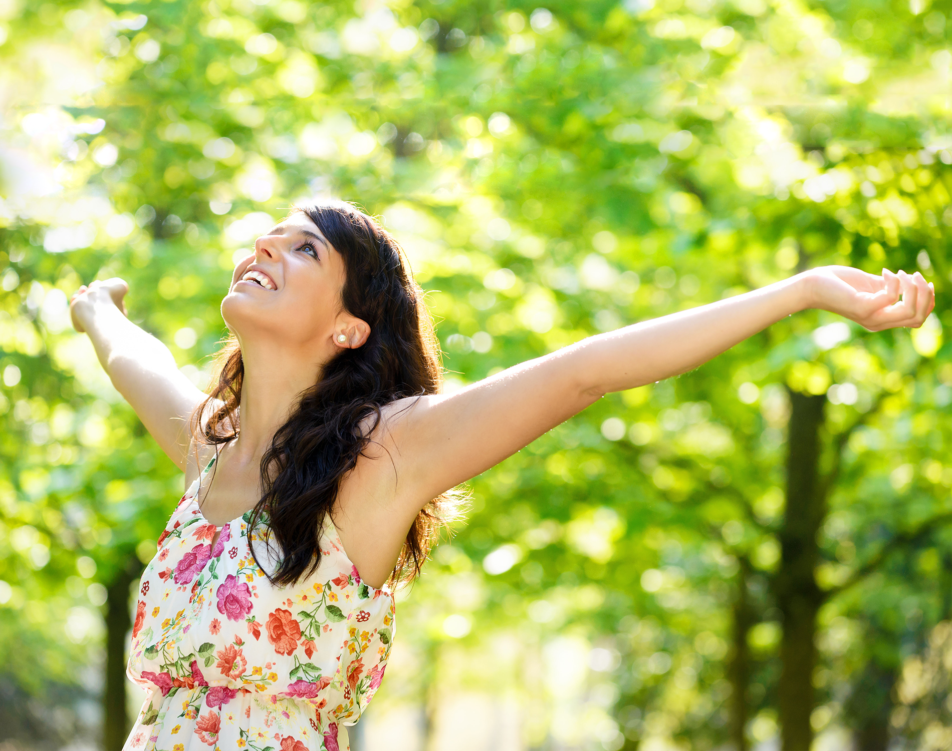 A woman in a floral dress is standing in a park with her arms outstretched.