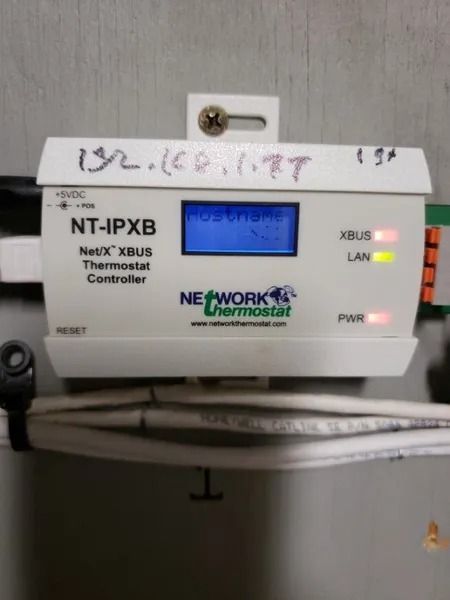 White Network Thermostat controller. Blue display, labeled “hostnam”.  XBUS & LAN lights, IP address noted.
