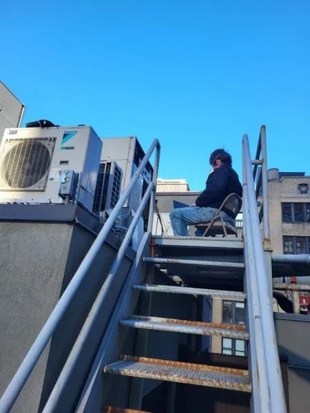 Person sits in a chair on a rooftop near air conditioning units. Blue sky.