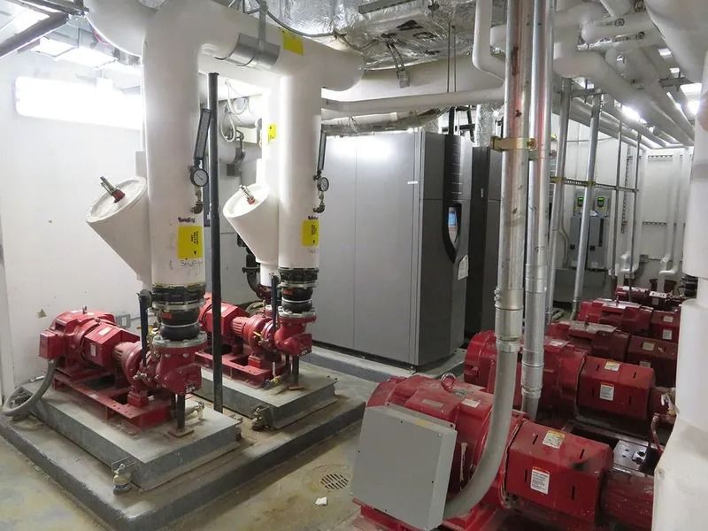 Pumps and piping in a mechanical room, red pumps on concrete, white pipes, gray metal machinery.