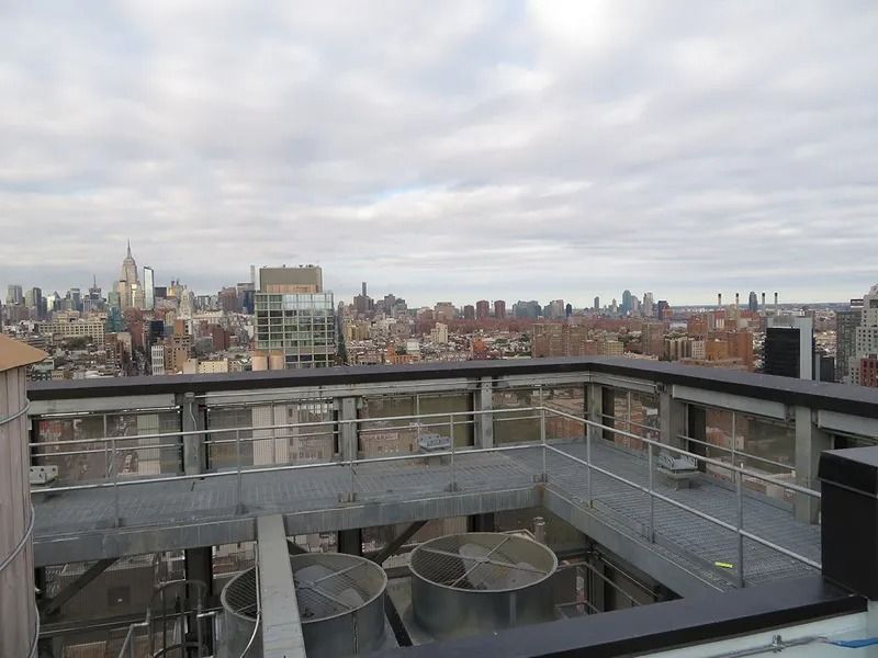 City skyline view from a rooftop. Steel platform with railings, mechanical equipment, and cloudy sky.