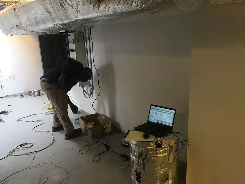 A person working on electrical equipment in a construction area, using a laptop, near wiring and ductwork.