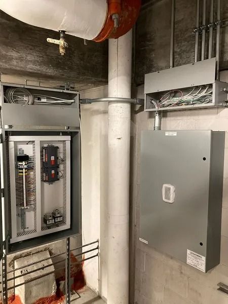 Electrical panels and conduit mounted on a concrete wall near a large pipe.