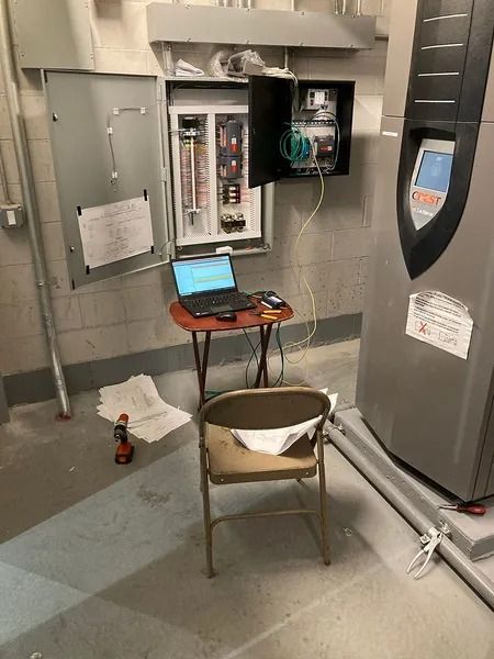 Electrical panel open with wires, laptop on a table, and a chair. Gray utility room.