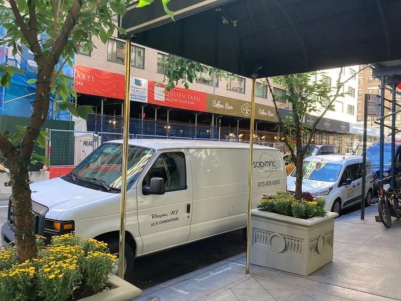White van parked under a canopy on a city street, yellow flowers in a planter, other vehicles visible.