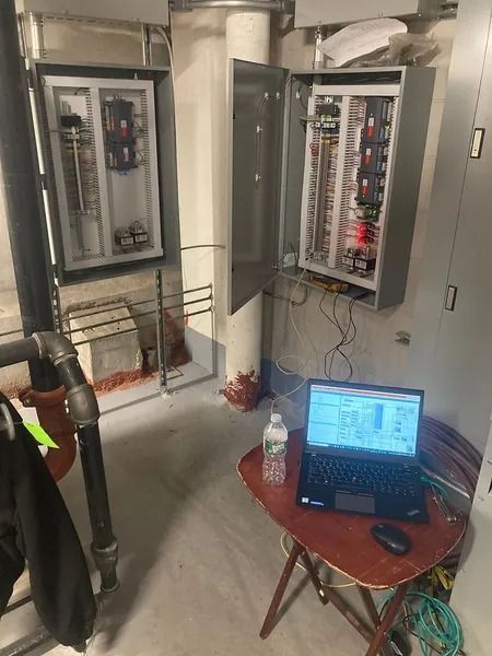 Electrical panels with open doors, a laptop on a table, and piping in an industrial setting.