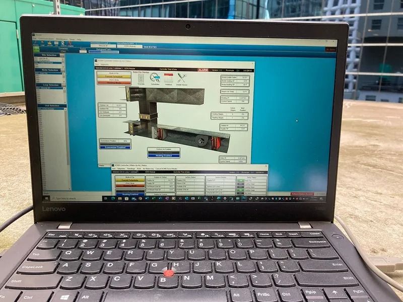 Laptop displaying a software interface outdoors, showing a mechanical device diagram. Blue sky and building visible.