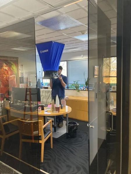 Man holding a blue ventilation device in an office. Glass walls reflect the interior; desk, chairs, and window visible.