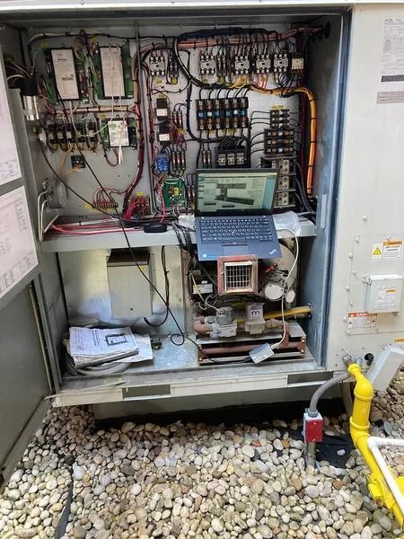 Open electrical panel with laptop, wires, and components. Yellow pipe visible outside.