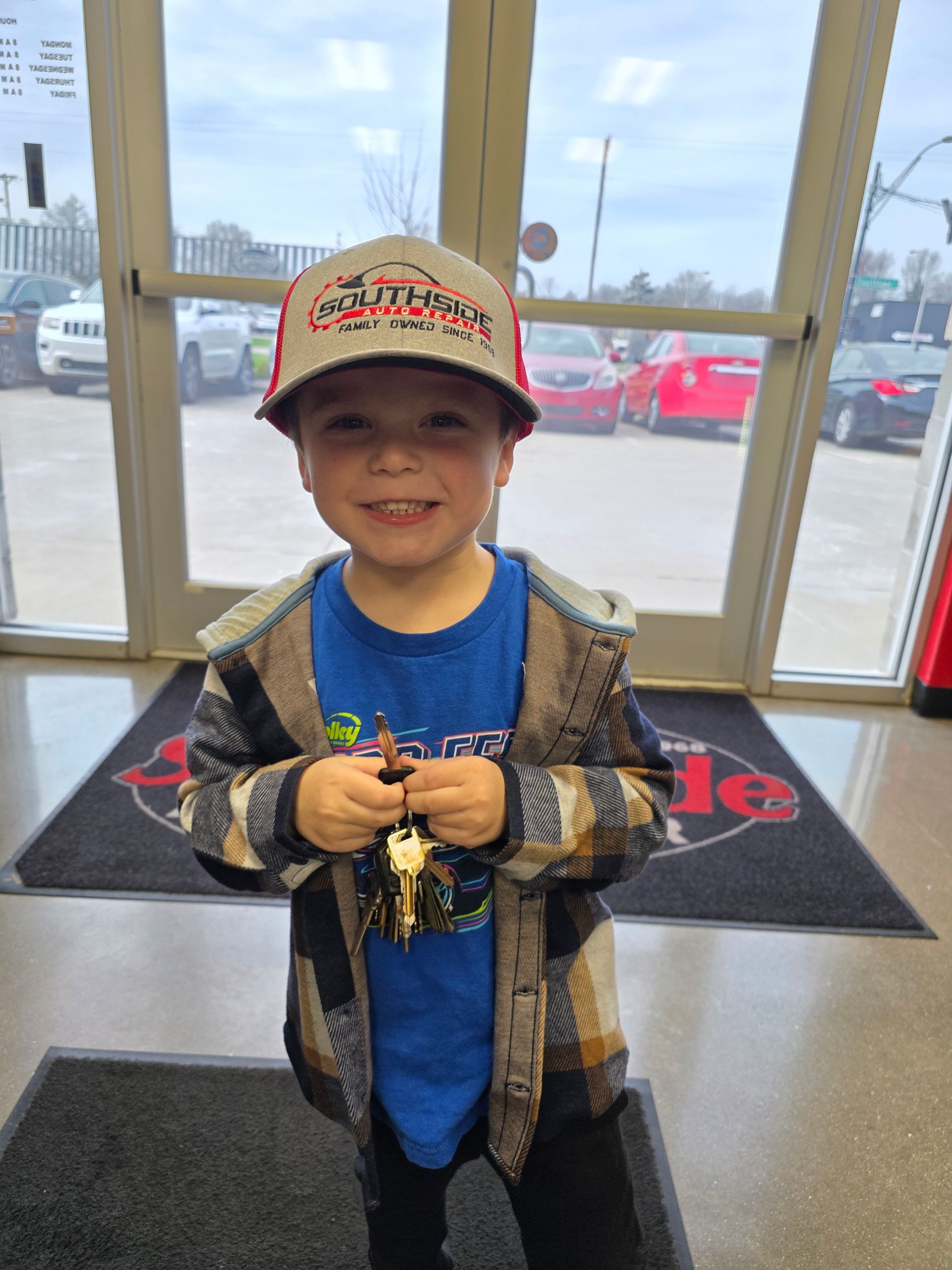 A smiling child in a hat and flannel shirt holding car keys while standing in a dealership doorway.