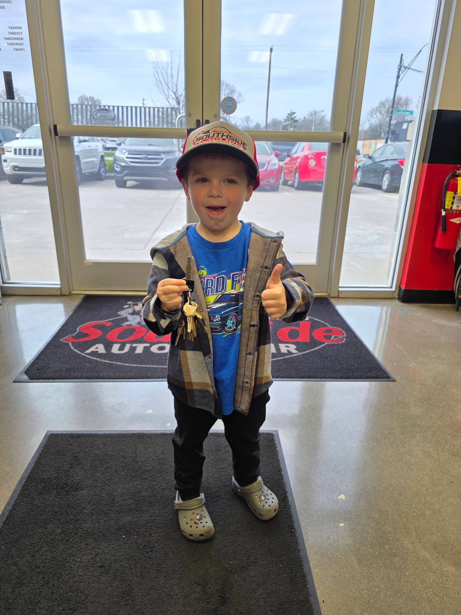 A child in a plaid jacket, blue shirt, and cap gives a thumbs up while holding car keys inside an auto dealership.