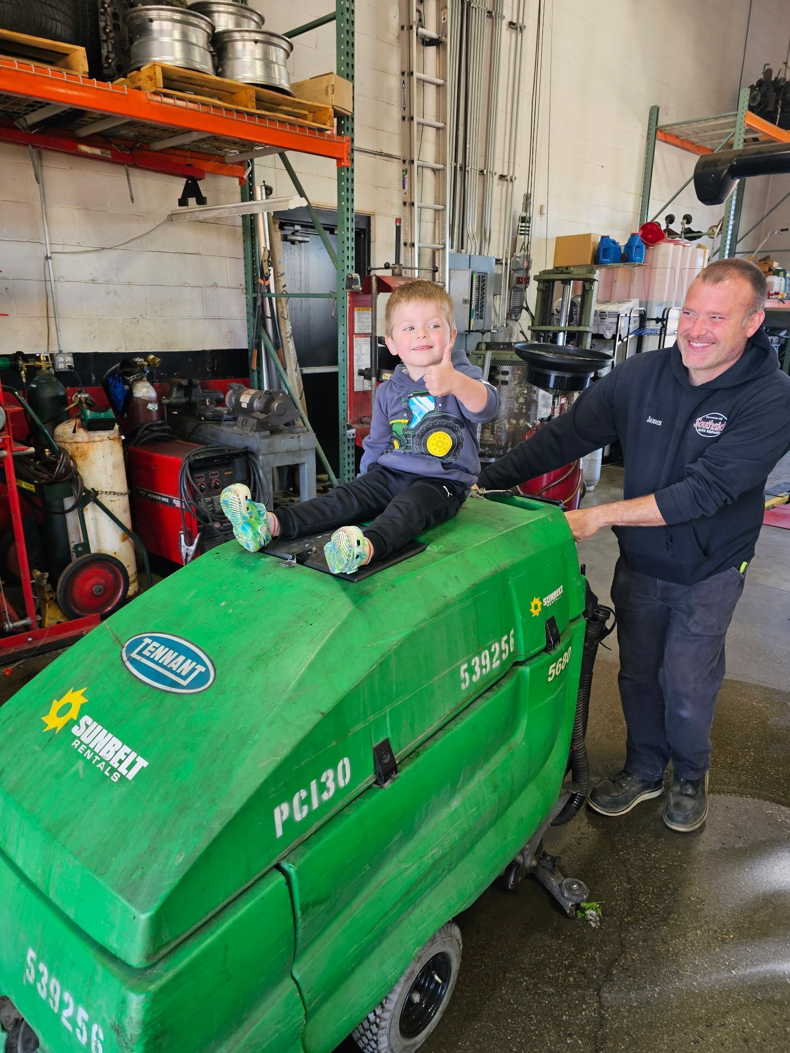 A child gives a thumbs up while sitting on a green industrial floor scrubber next to a smiling person in a workshop.