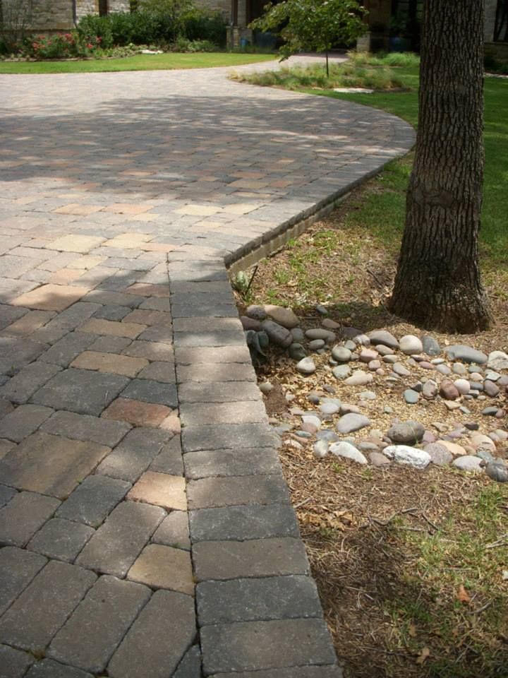 A brick walkway leading to a driveway with a tree in the background.