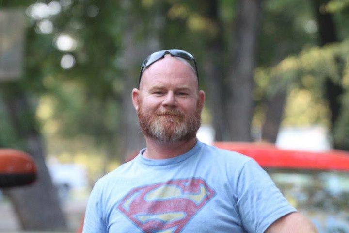 A man with a beard and sunglasses is wearing a blue Superman shirt.