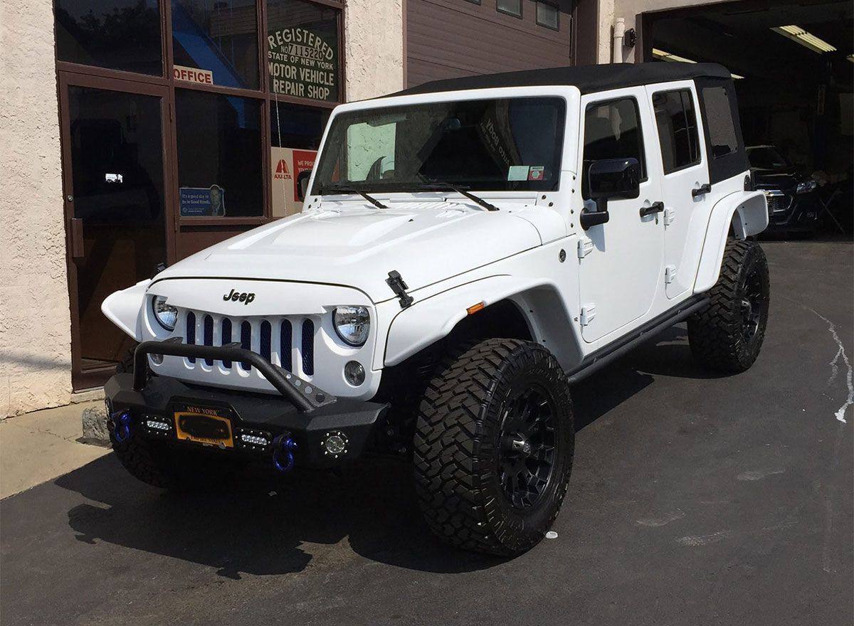 White Jeep Wrangler with large off-road tires parked on a street near buildings.