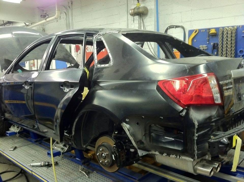 Damaged black sedan on a tow lift inside an auto repair shop, with a wheel removed.