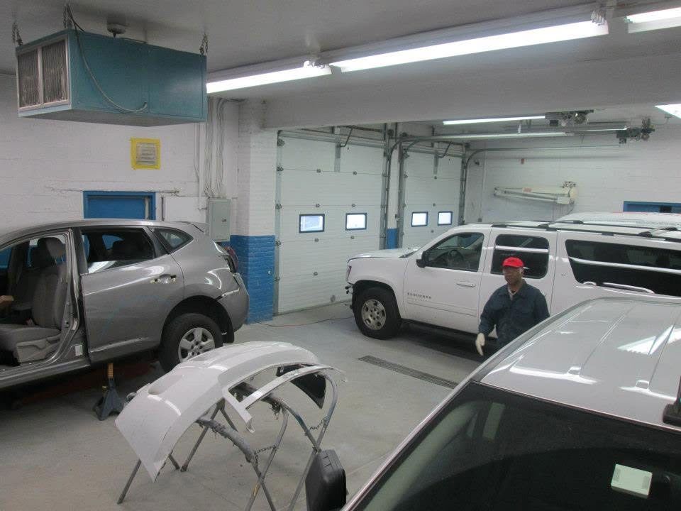 Auto body shop with two vehicles, car parts on the floor, and a person in a red cap near a white van