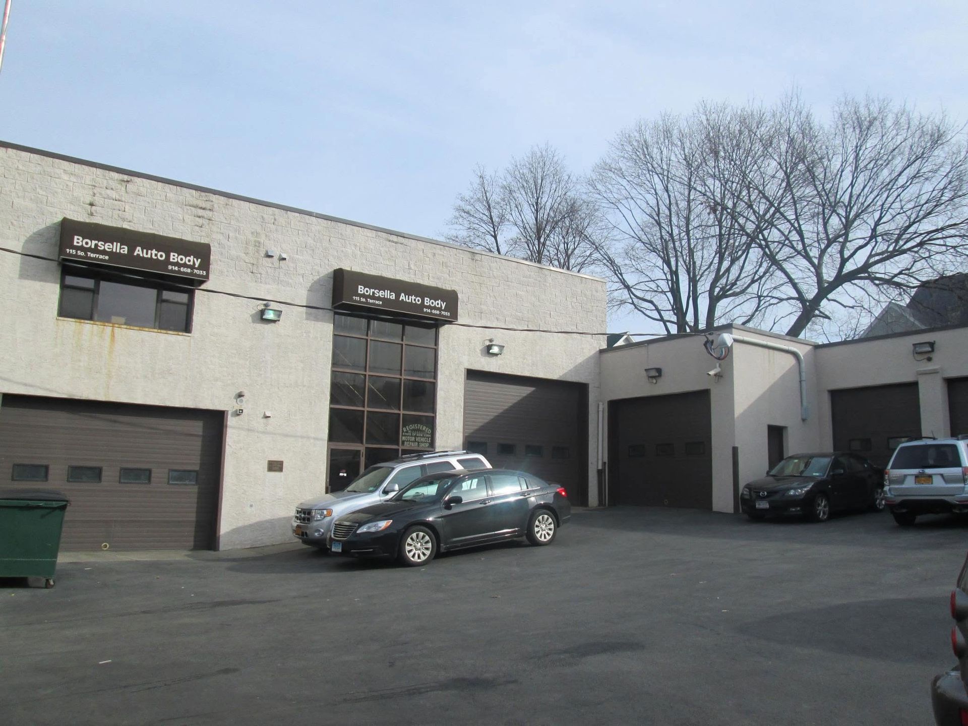 Industrial building with garage doors and parked cars in a paved lot under bare trees