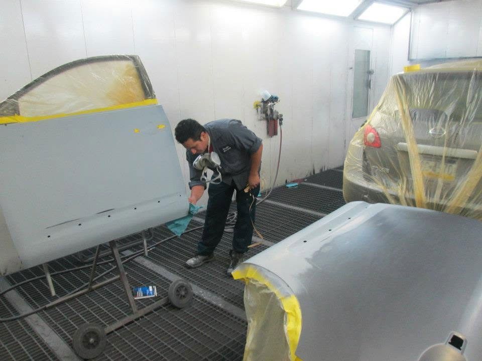 Worker painting car parts in a spray booth, with masked vehicle panels and tools on the floor.