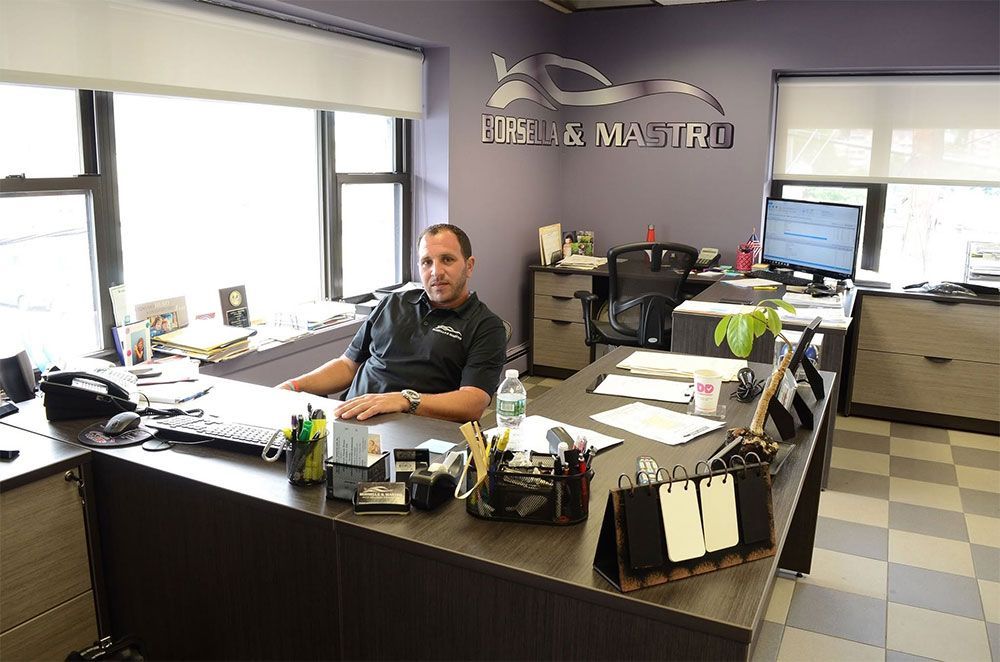 Office interior with a man seated at a cluttered desk near windows and computer monitors.