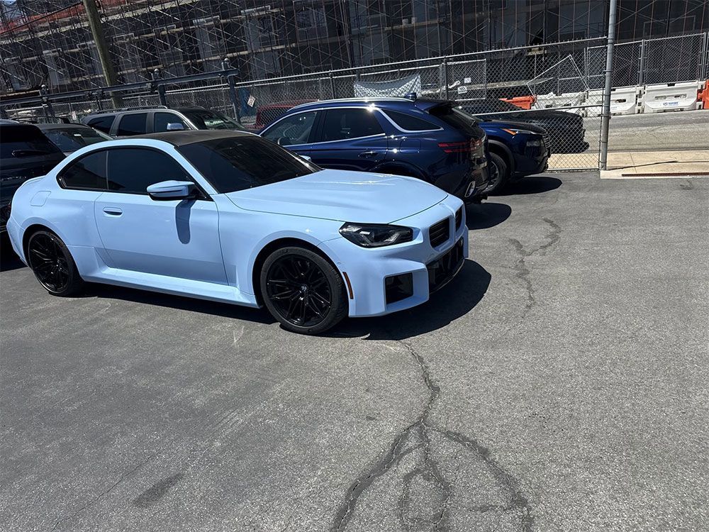 White sports sedan parked on asphalt in front of a chain-link fence, with dark wheels and a blue SUV behind it