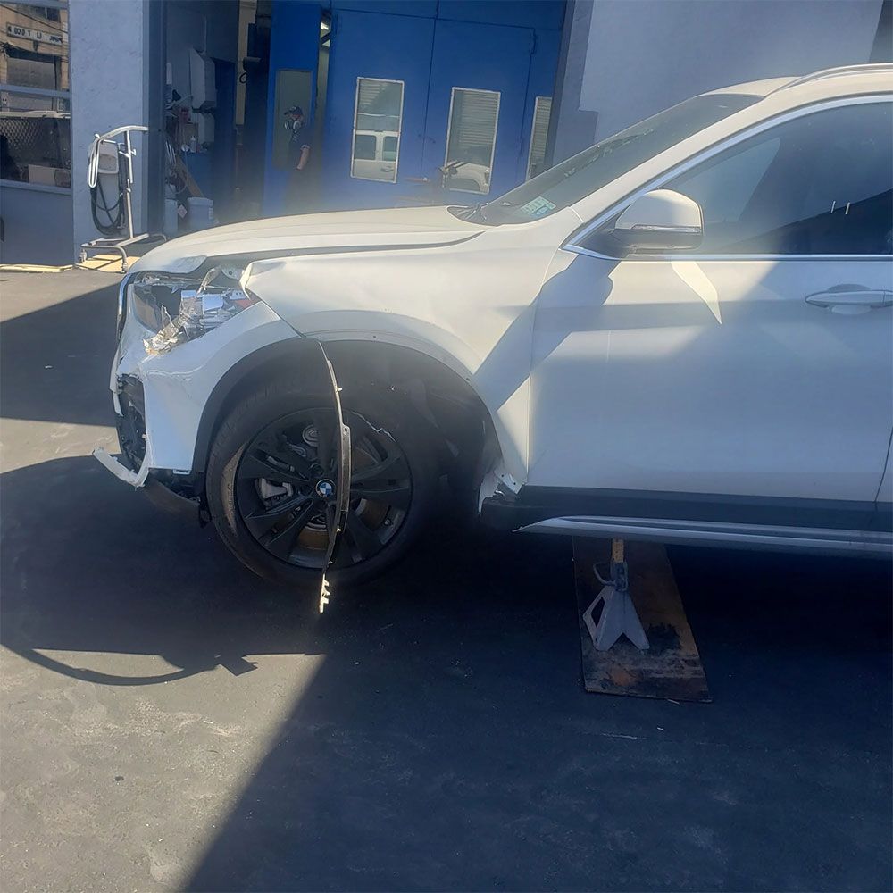 Damaged white SUV with front-left wheel off, supported by a jack in a driveway after a collision