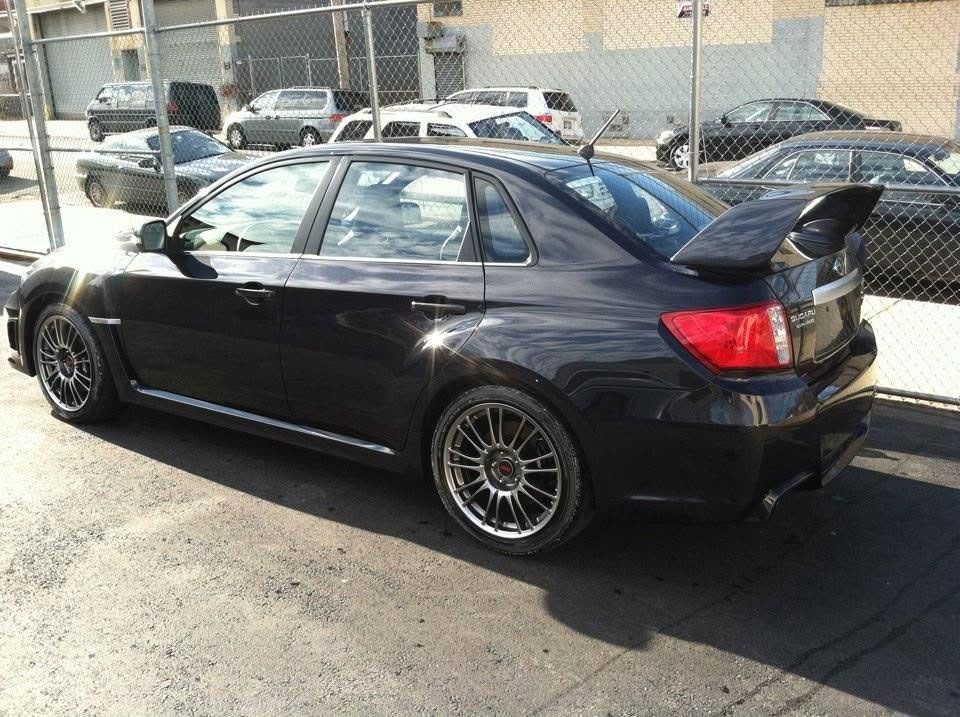 Black sedan parked in a lot, side view with large rear spoiler and chain-link fence behind it