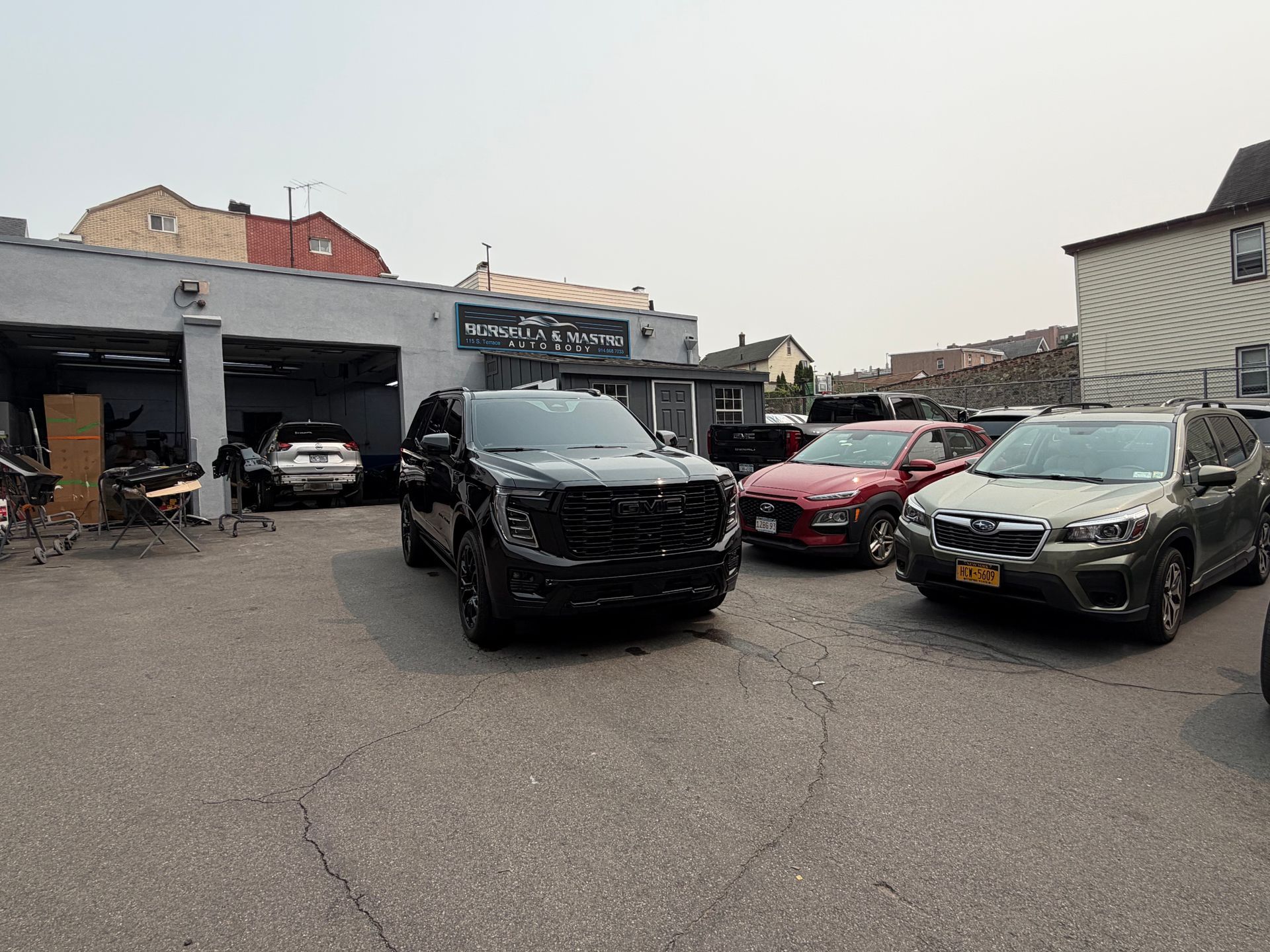 Parking lot with several cars, including a black SUV and red sedan, beside auto shops on an overcast day