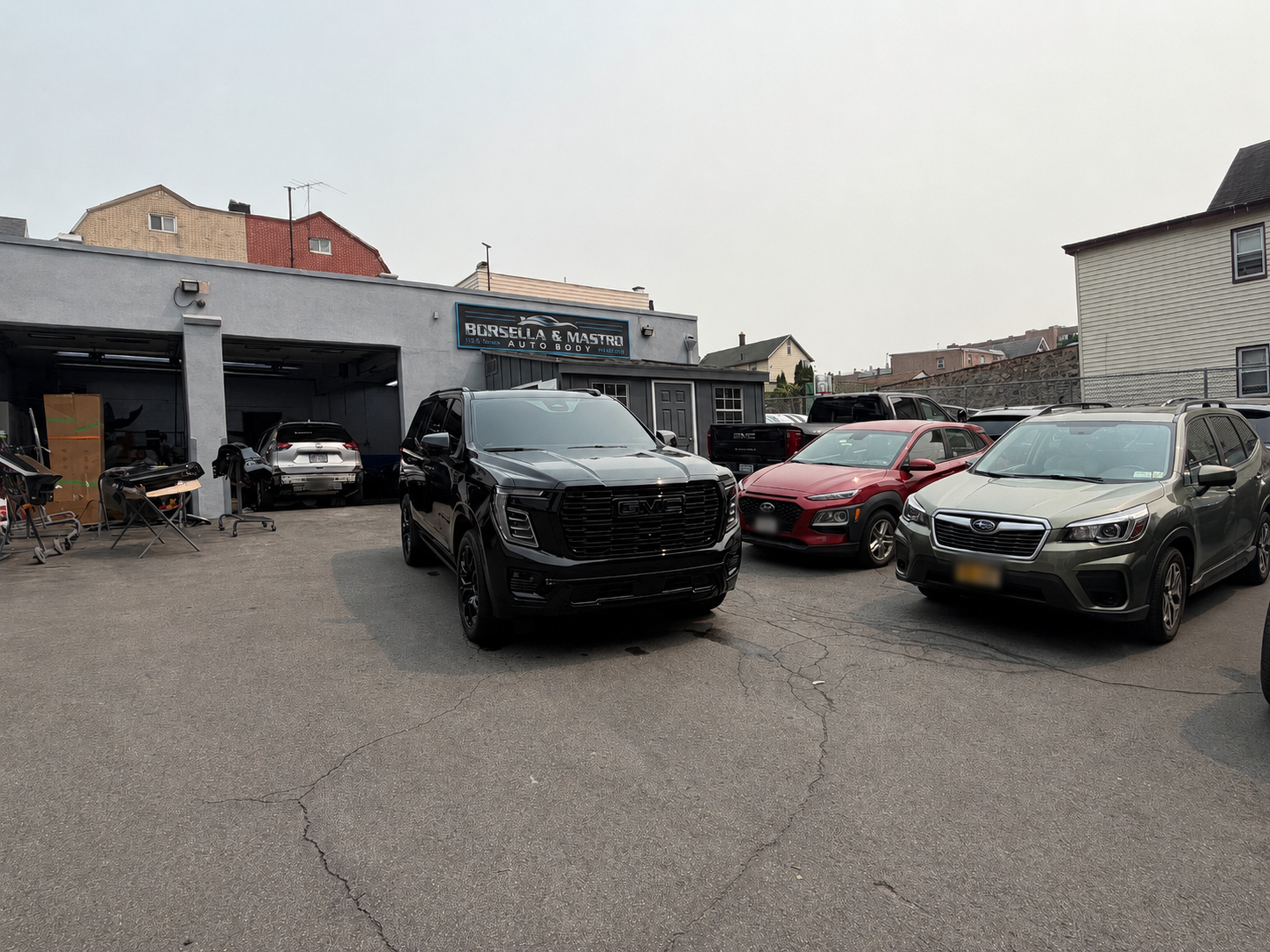 Parking lot with several cars, including a black SUV in front of a garage under an overcast sky.