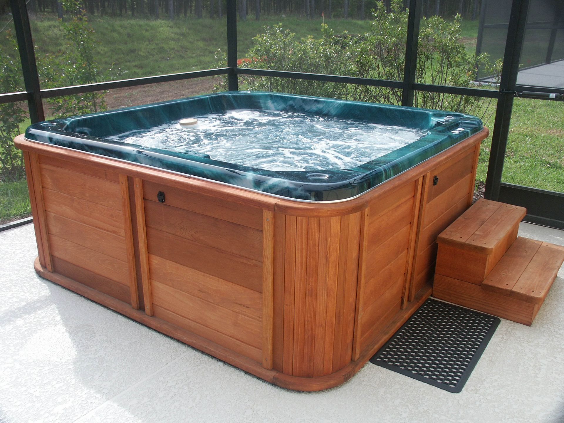 A square hot tub with wood paneling and a matching step stool, situated on a patio inside a screened enclosure.