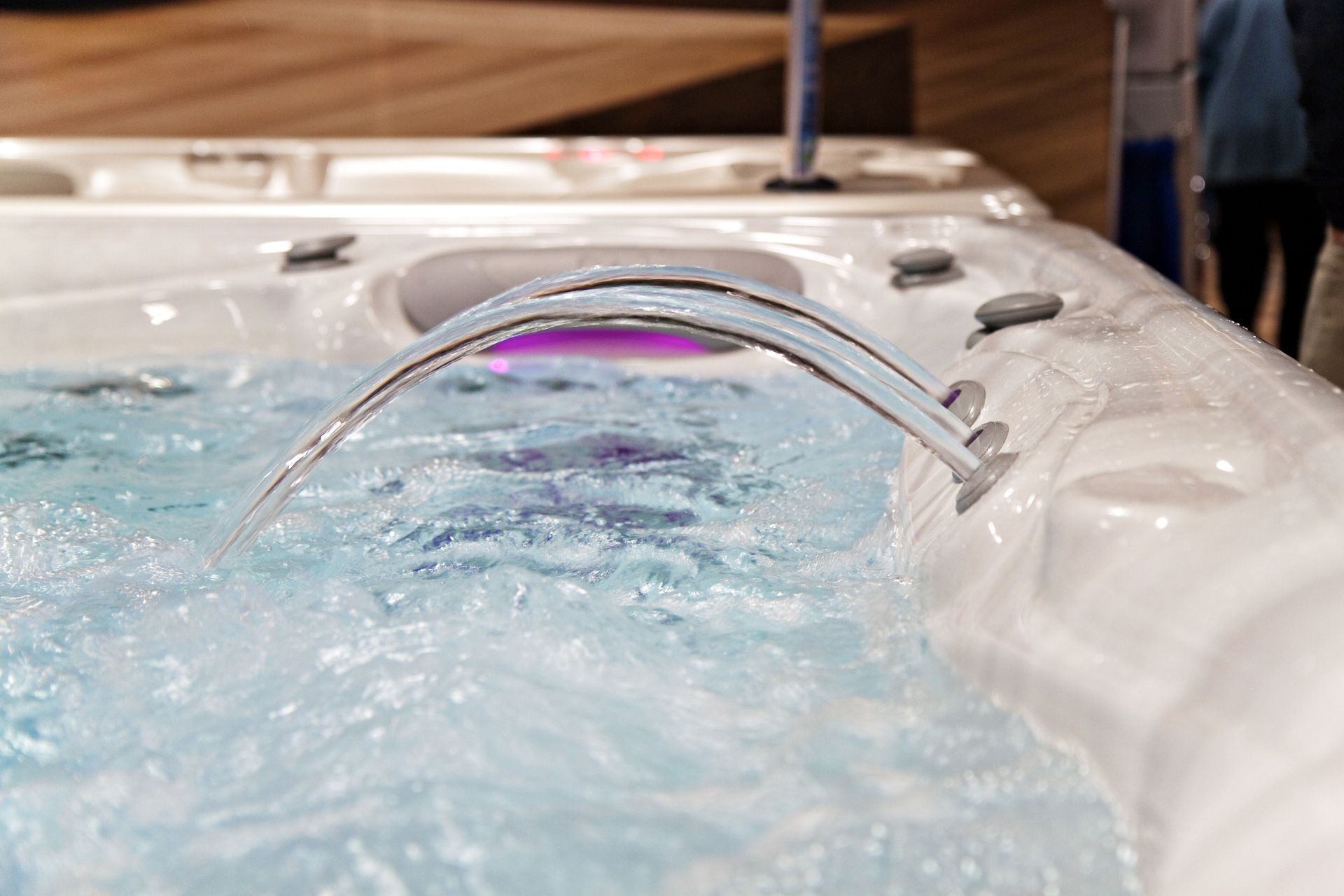 A gentle arc of water flows from a sleek faucet into the bubbling, illuminated turquoise water of a white hot tub.