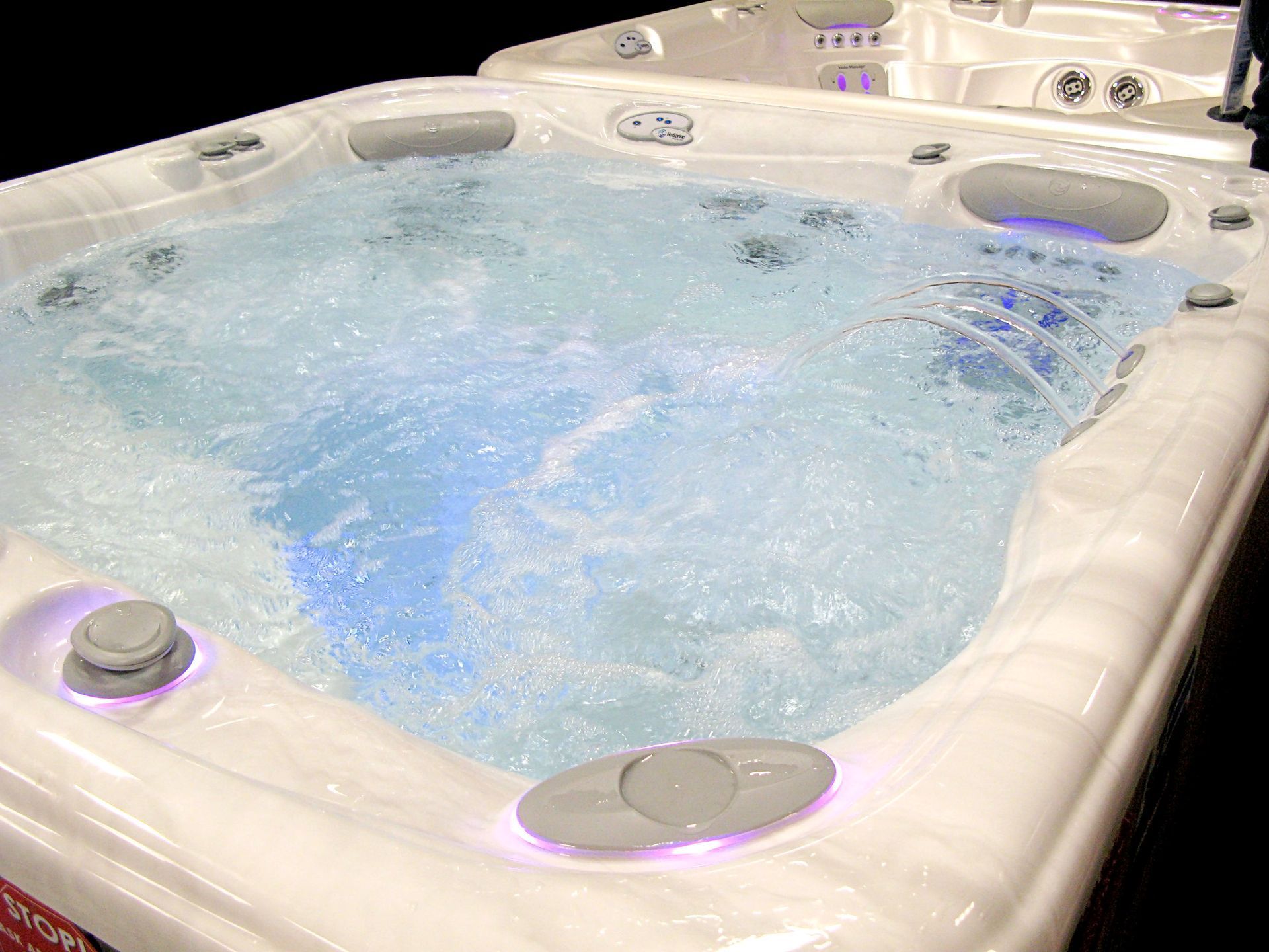 A white, square hot tub filled with bubbling, illuminated blue water, featuring molded seats and headrests.