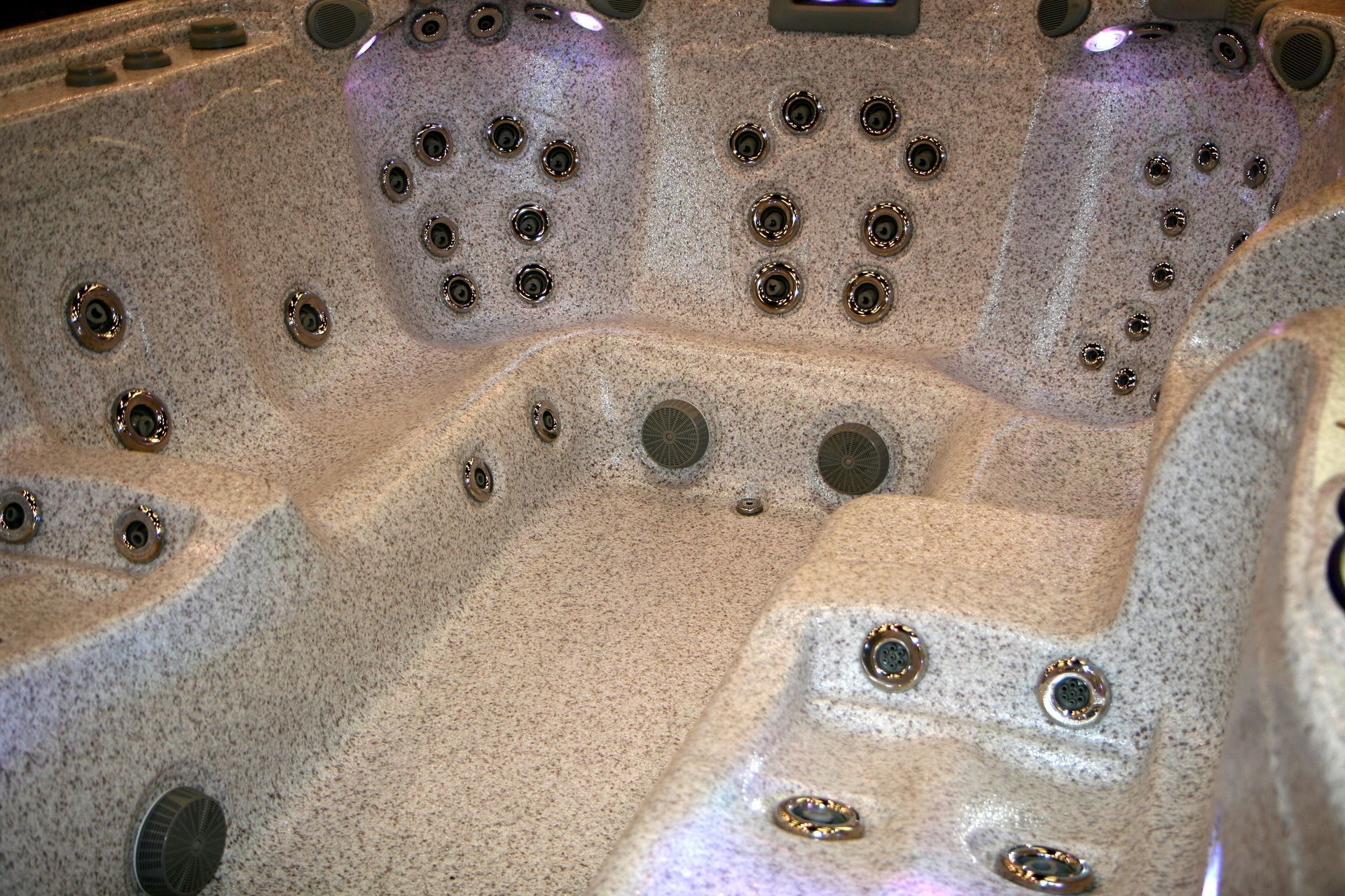 An interior view of a textured, beige hot tub with multiple jet nozzles arranged on the seats and walls.