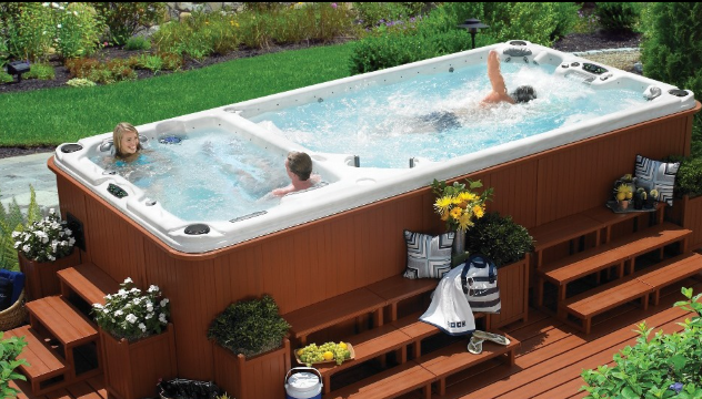 A high-angle view of a rectangular dual-zone swim spa on a wooden deck, with three people relaxing and swimming inside.