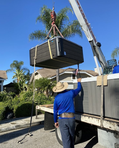 A crane lifts a large, dark-colored hot tub off a flatbed truck while a person guides it outdoors on a sunny day.