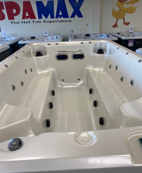 A bright white, rectangular swim spa on display in a showroom with the SPAMAX logo on the wall in the background.