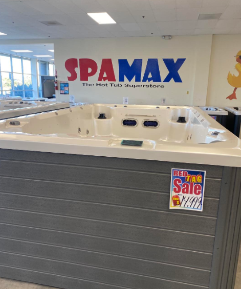 A white hot tub sits in a showroom labeled SPA MAX with a Red Tag sale sign displayed on its gray paneled side.