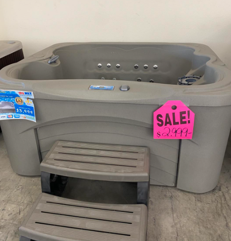 A gray, square hot tub with matching steps on a sale display, featuring a bright pink sale sign marked $2,999.
