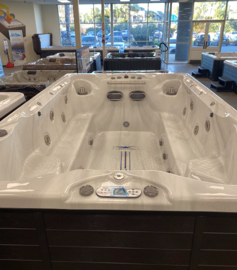 A white rectangular swim spa with a dark brown exterior, displayed inside a showroom with large glass windows.