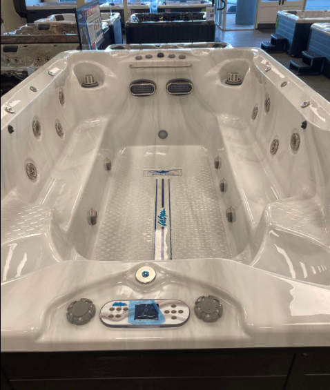 An empty, white rectangular swim spa with integrated jets and a digital control panel, displayed in a showroom.