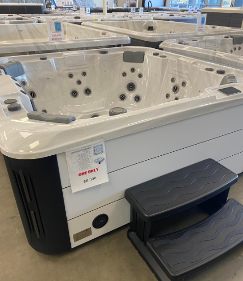 A white hot tub with a black corner panel and matching steps displayed in a showroom with other units.