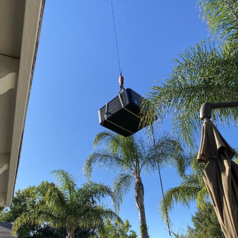 A large, dark, rectangular object hangs from a crane line against a bright blue sky, with palm trees visible below.