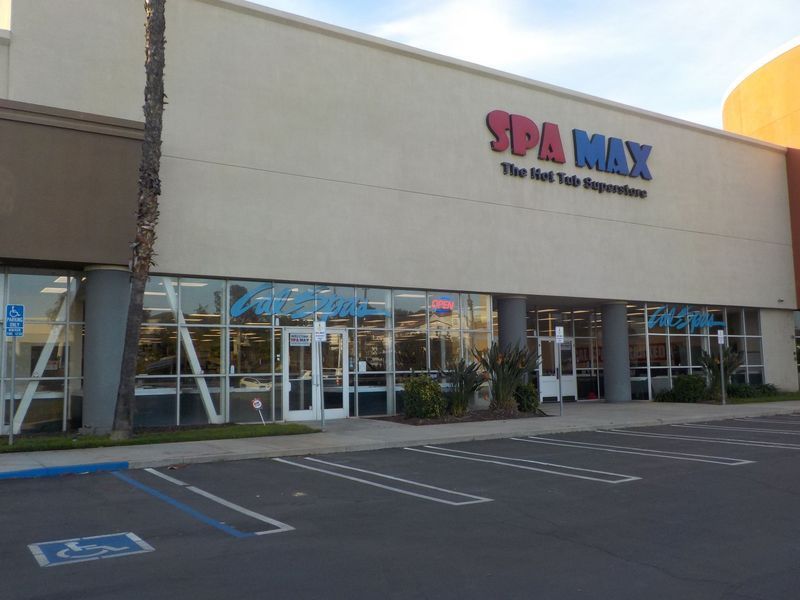 Exterior of Spa Max, a hot tub store with blue lettering and large glass windows, viewed from a parking lot.