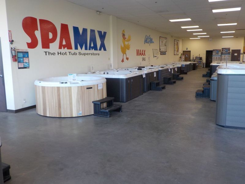 A showroom interior with several hot tubs lined up on a concrete floor beneath the large SpaMax logo on the wall.