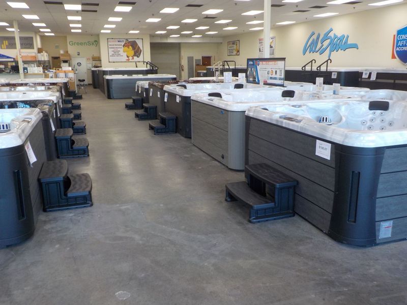 A showroom with rows of grey hot tubs, each with matching black steps, arranged on a concrete floor under bright lights.